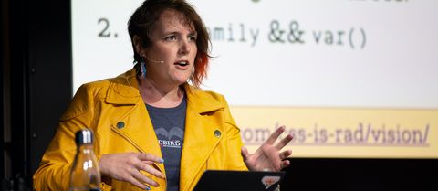 Miriam on stage talking and gesturing
in front of a projection screen
wearing a yellow leather jacket
and white-blue-pink lightning-bolt earrings