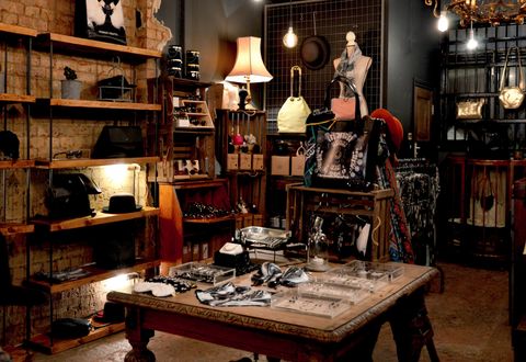 A dark vintage accessory store lit by lamps and bare bulbs, with bags, jewelry, sunglasses and a bowler hat on the wooden shelves and carved table.
